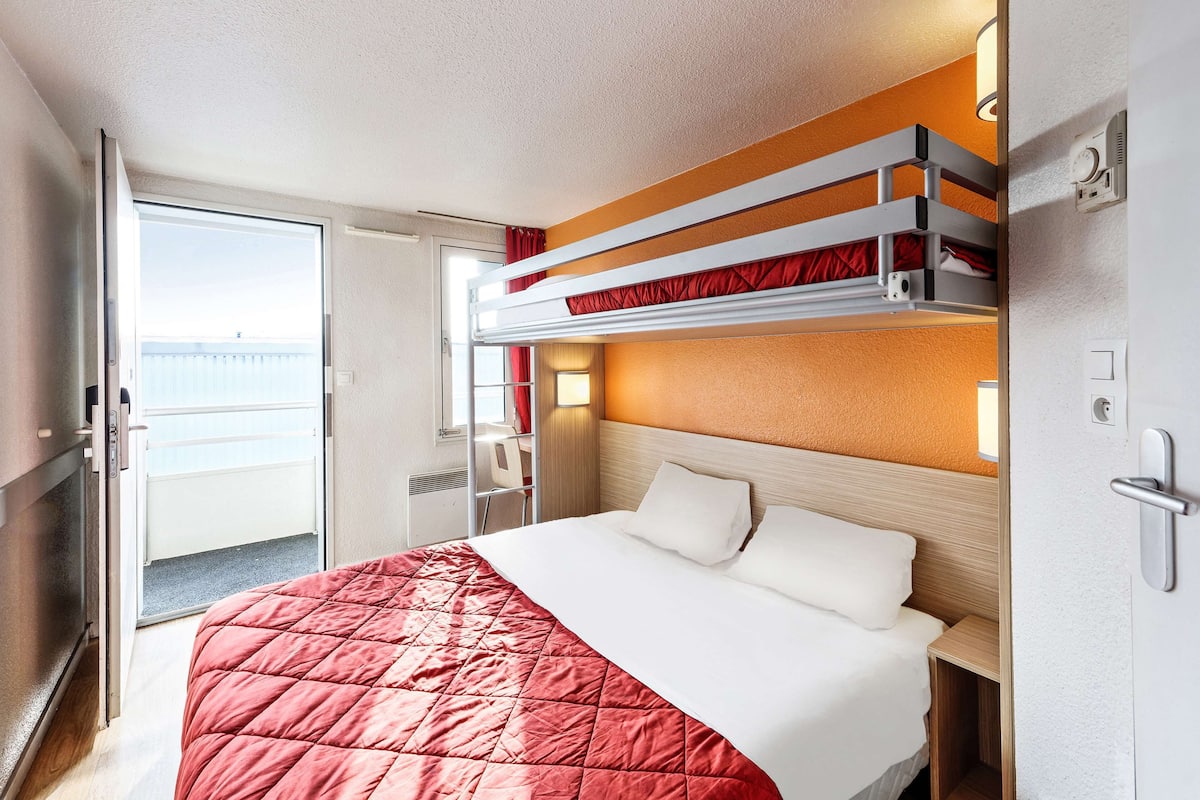 A hotel room with a bunk bed, a separate bed, a desk, and a view of the sea.