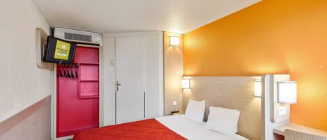 A hotel room with a bed, a red bedspread, a TV mounted on a wall, a wardrobe, and a nightstand with a lamp.