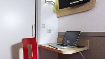 Une chambre d’hôtel compacte, dotée d’un bureau en bois, d’une chaise rouge, d’un ordinateur portable, d’un téléviseur à écran plat fixé au mur et d’une petite table de chevet.