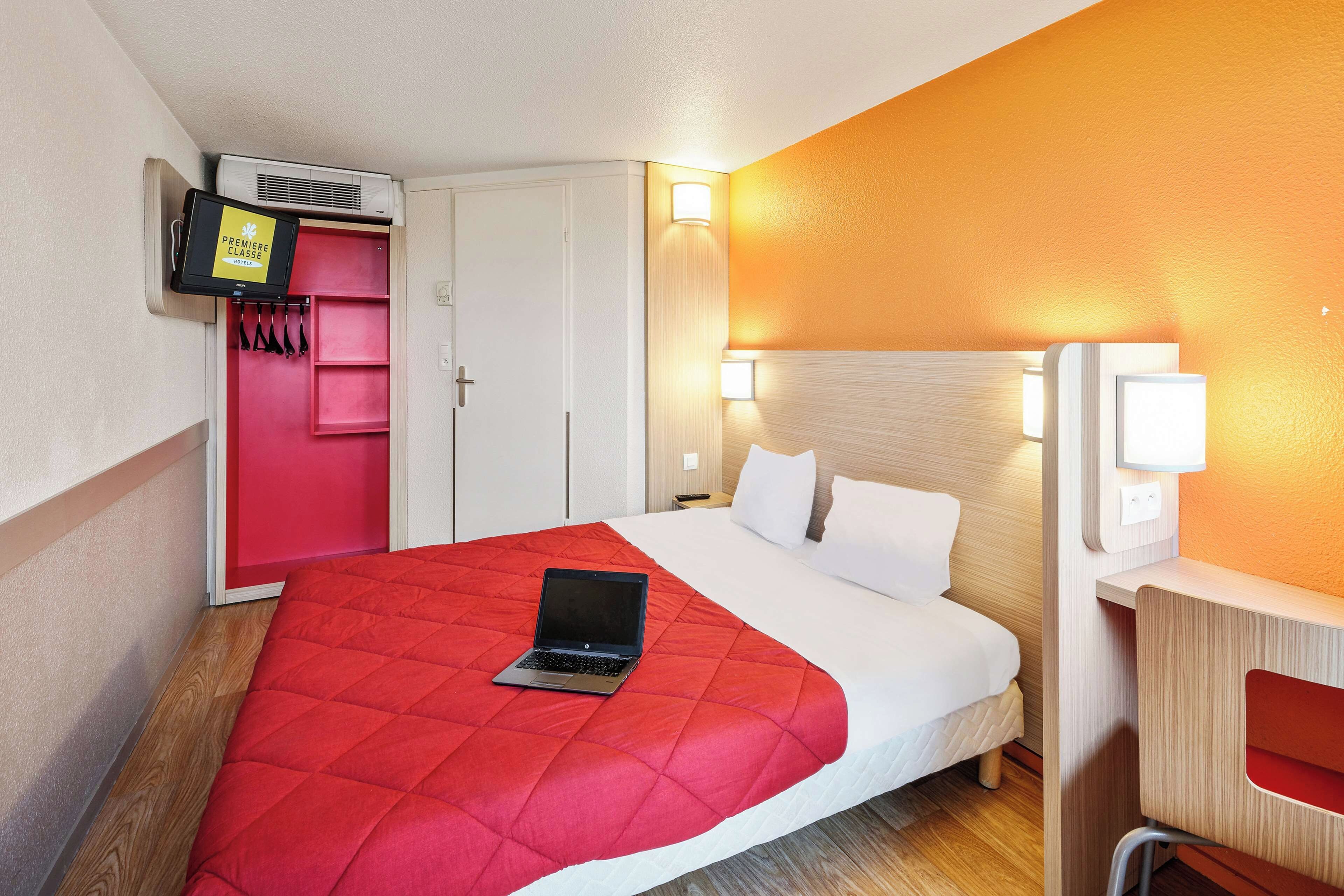 Habitación de hotel con cama, laptop sobre la cama, armario, escritorio con lámpara y aire acondicionado de pared.