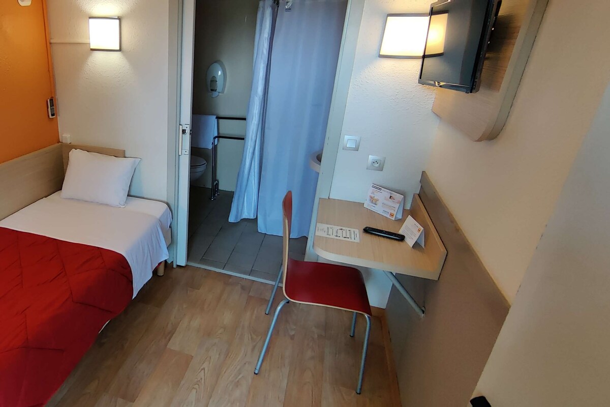 Una habitación de hotel pequeña con dos camas, un escritorio, una silla y un rincón con ducha.