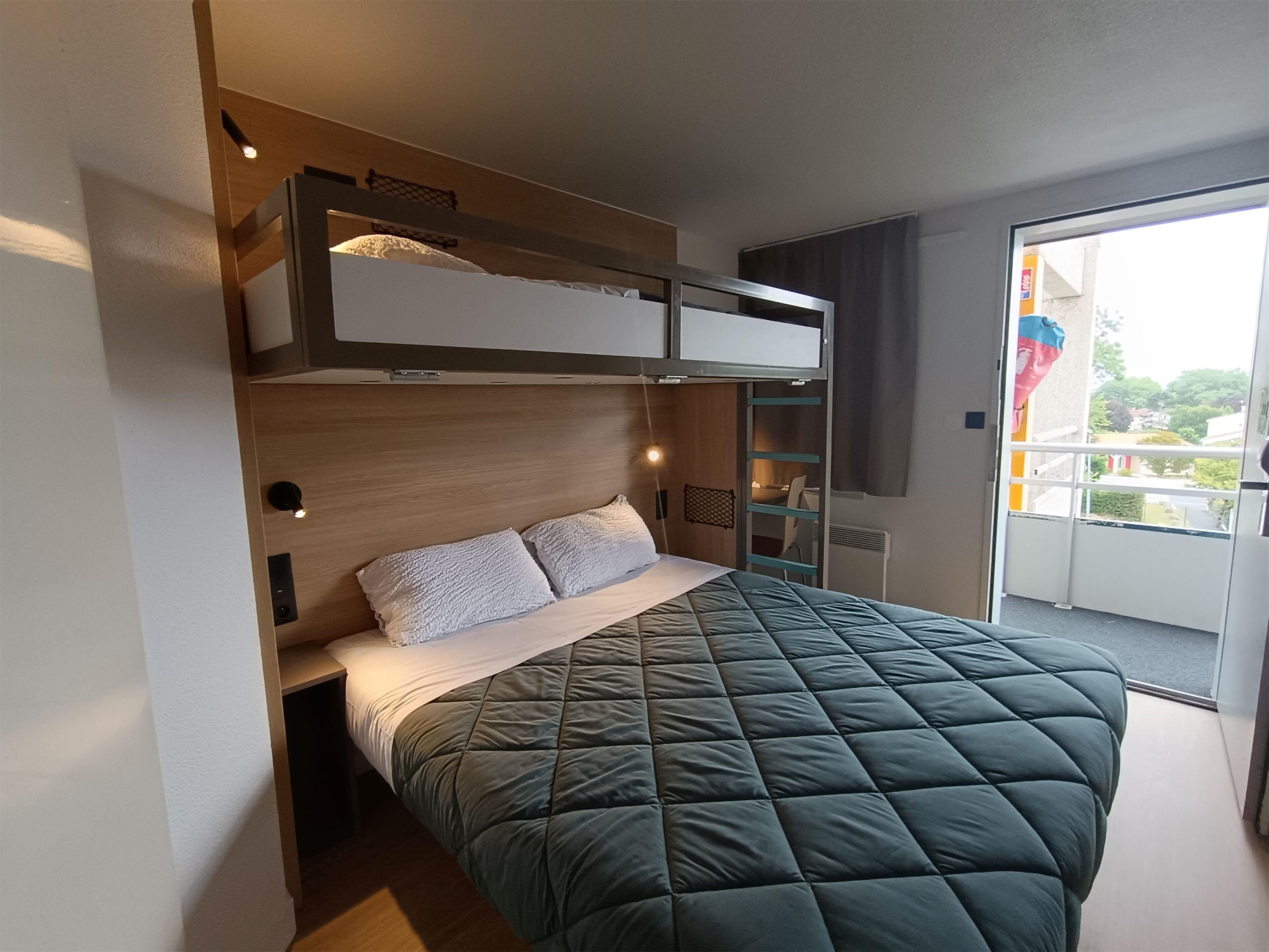 A modern hotel room with a bunk bed, a large bed with a quilted green duvet, a wooden headboard, and a sliding door leading to a balcony.