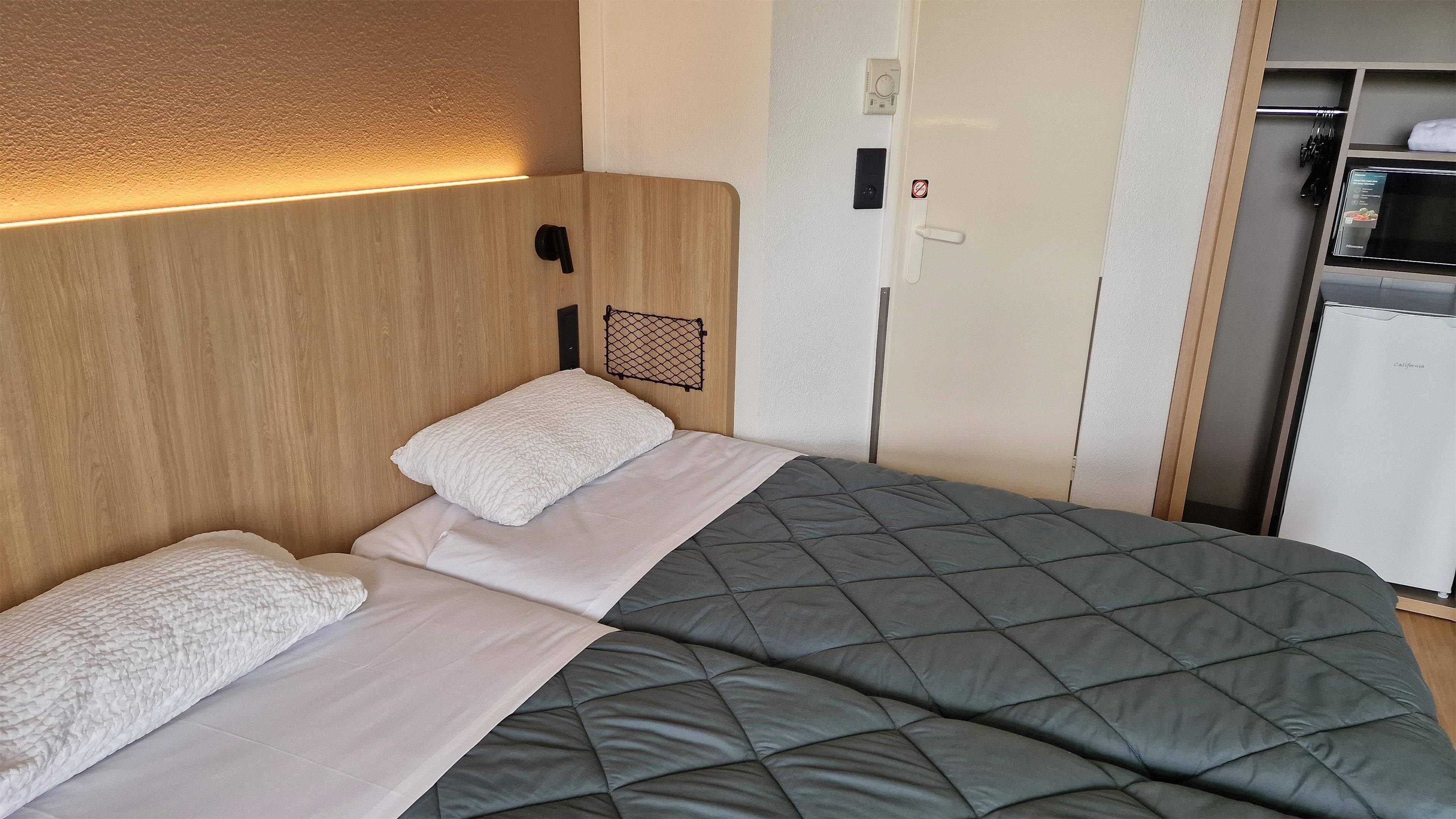 A neatly made bed with a grey quilt and white pillows, a wooden headboard, a small shelf with a black object, and a white door with a 'no smoking' sign.