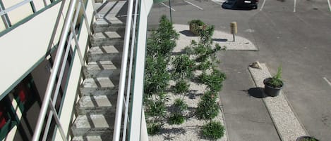 Entrada de un hotel con una escalera que lleva al vestíbulo, un aparcamiento con varios coches y un pequeño jardín con plantas en macetas.
