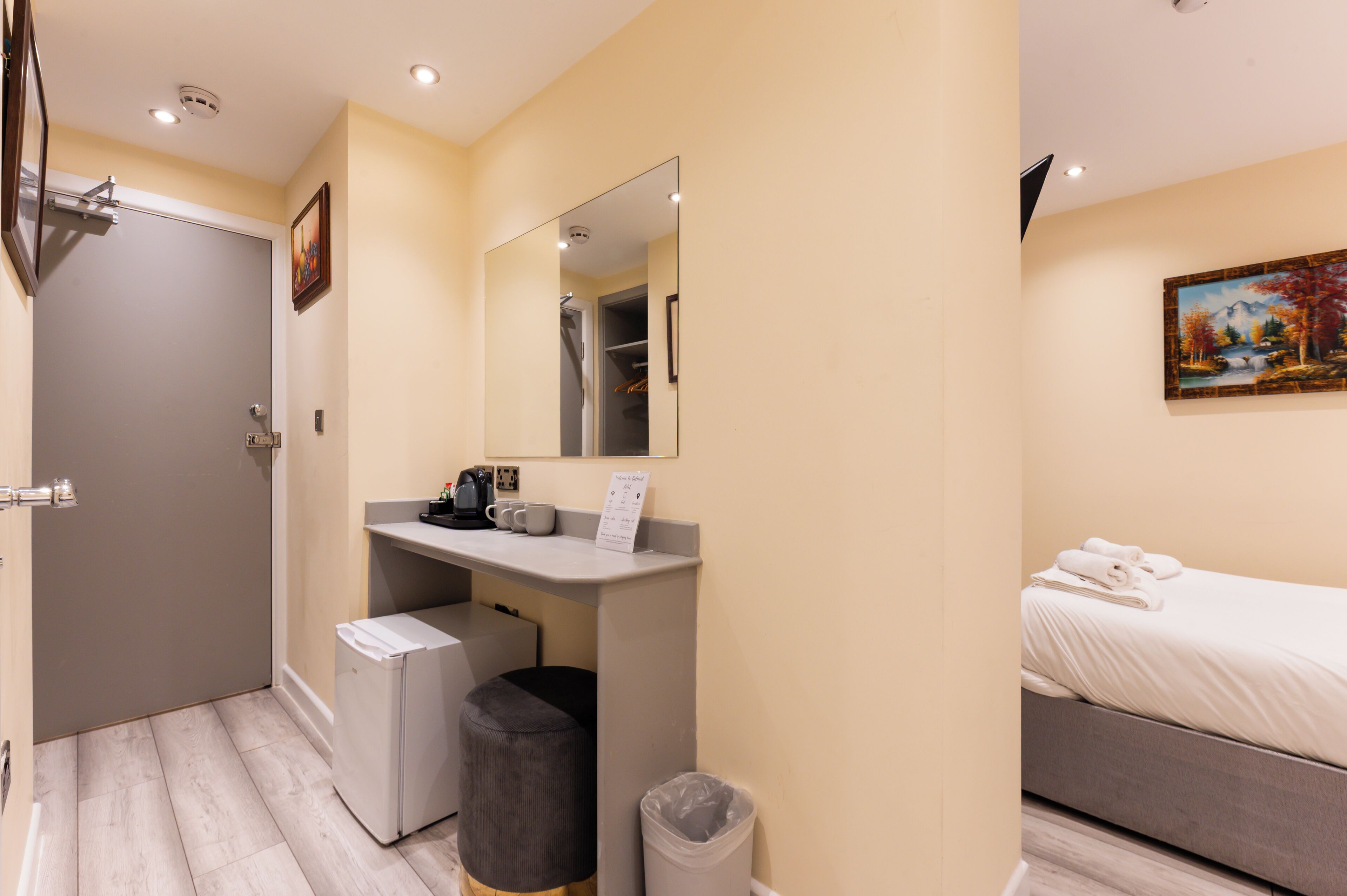 A compact hotel room with a bed, a small desk with a mirror, a trash bin, and a painting on the wall.