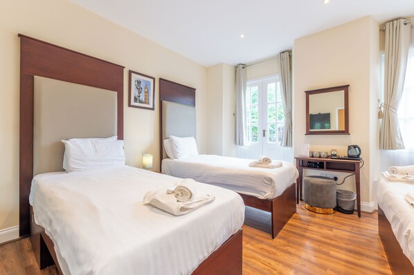 A hotel room with two beds, a desk with a chair, a small stool, a trash bin, and a window with curtains.