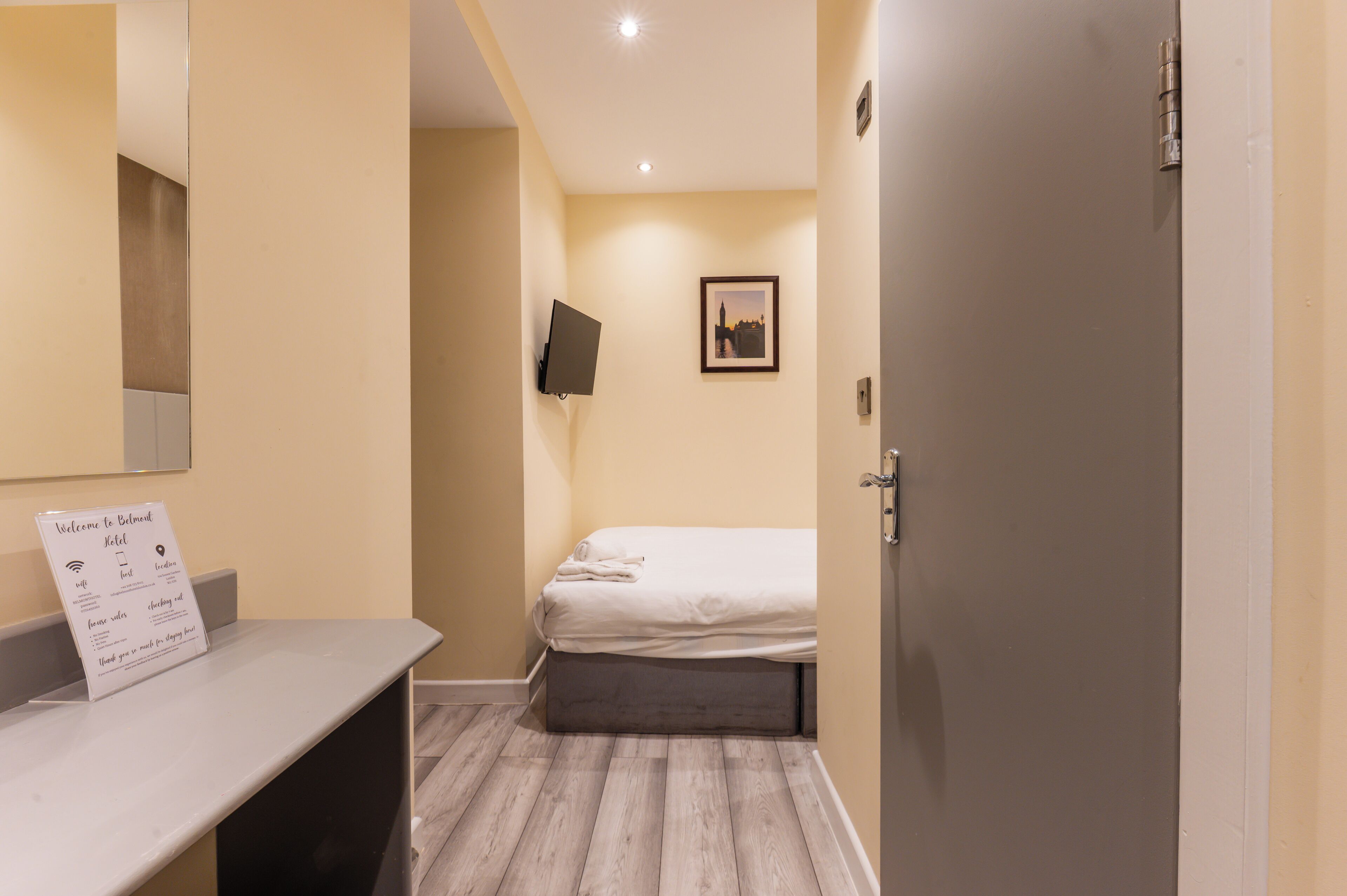 A hotel room with a bed, a flat-screen TV mounted on the wall, a small framed picture, and a welcome sign on the counter.