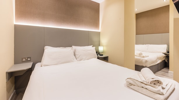 A modern hotel room with a large bed, a bedside table with a lamp, and a mirror reflecting the room. - Belmont Hotel (London)