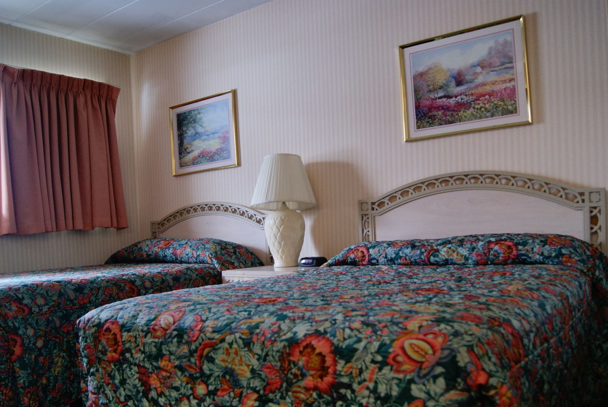 A hotel room with two beds, a nightstand, a lamp, and two framed pictures on the wall.