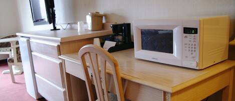 Una kitchenette pequeña con microondas, cafetera y un monitor de computadora sobre un escritorio.