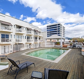 A rooftop pool area with lounge chairs and a covered seating area, surrounded by multi-story buildings with balconies.