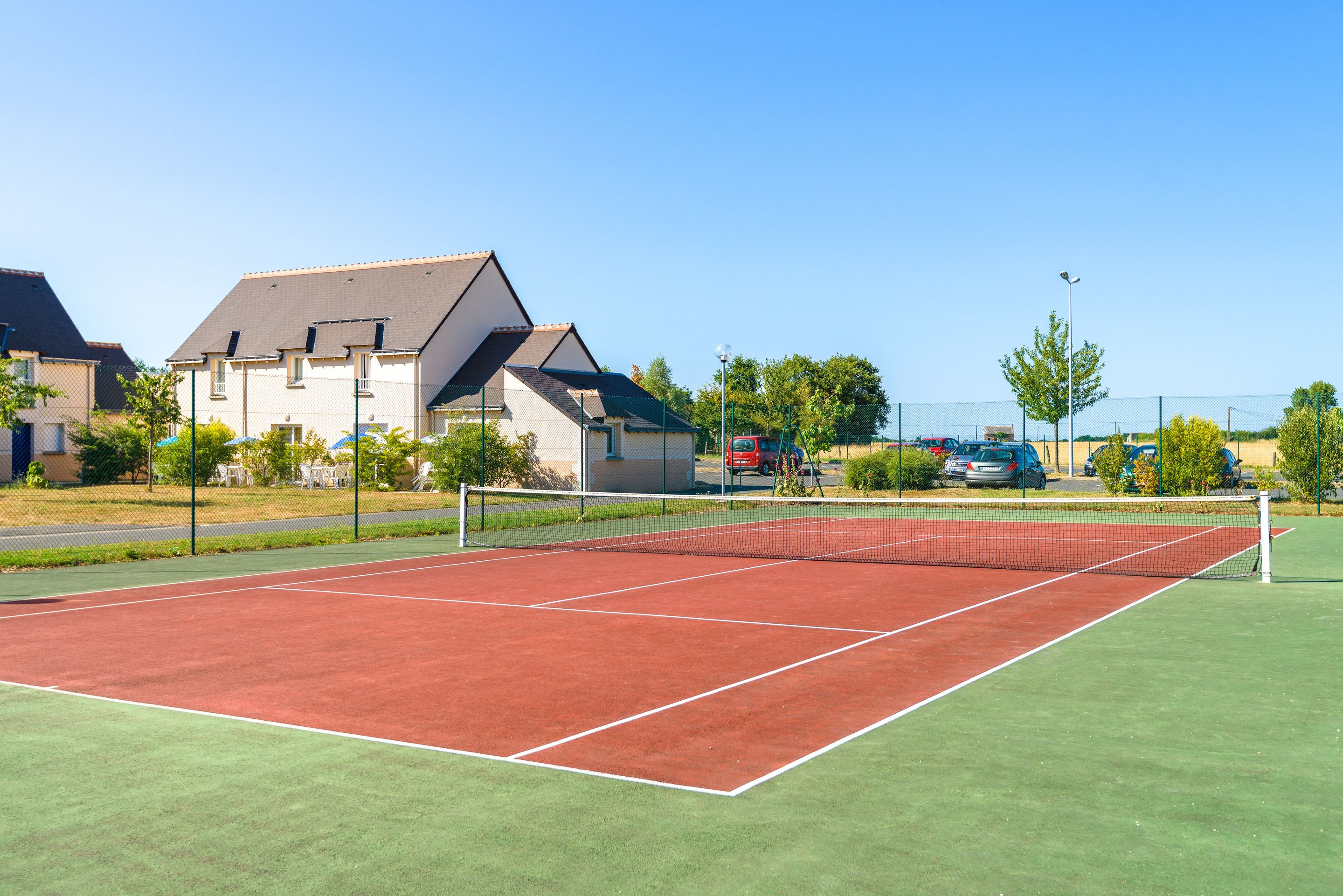 A tennis court with a net, surrounded by a fence, and houses in the background.