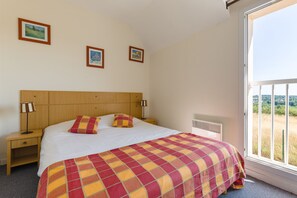 A bedroom with a wooden headboard, a checkered bedspread, two bedside lamps, and a window with a view of a field. - Résidence Lagrange Vacances Les Jardins Renaissance (Azay-le-Rideau)