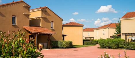Un quartier résidentiel où les maisons se distinguent par leurs toits en tuiles de terre cuite et leurs murs de couleur beige, ainsi qu’un chemin pavé et une végétation luxuriante.