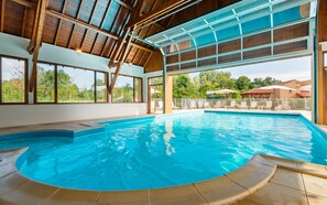 Una amplia piscina cubierta con agua azul cristalina, vigas de madera en el techo y grandes ventanales que ofrecen vistas de la vegetación circundante.