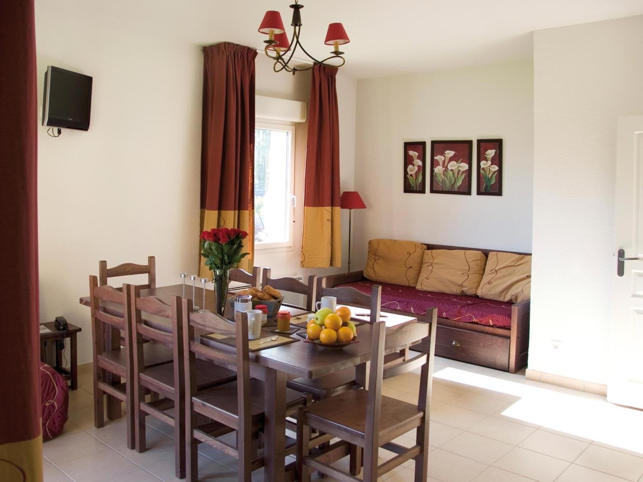 A dining area with a wooden table and chairs, a vase with flowers, and a bowl of fruit. There is a sofa and a wall-mounted TV in the background.