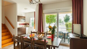 Uma sala de jantar com uma mesa de madeira posta para quatro pessoas, uma cozinha ao fundo e uma varanda com vista para o exterior.