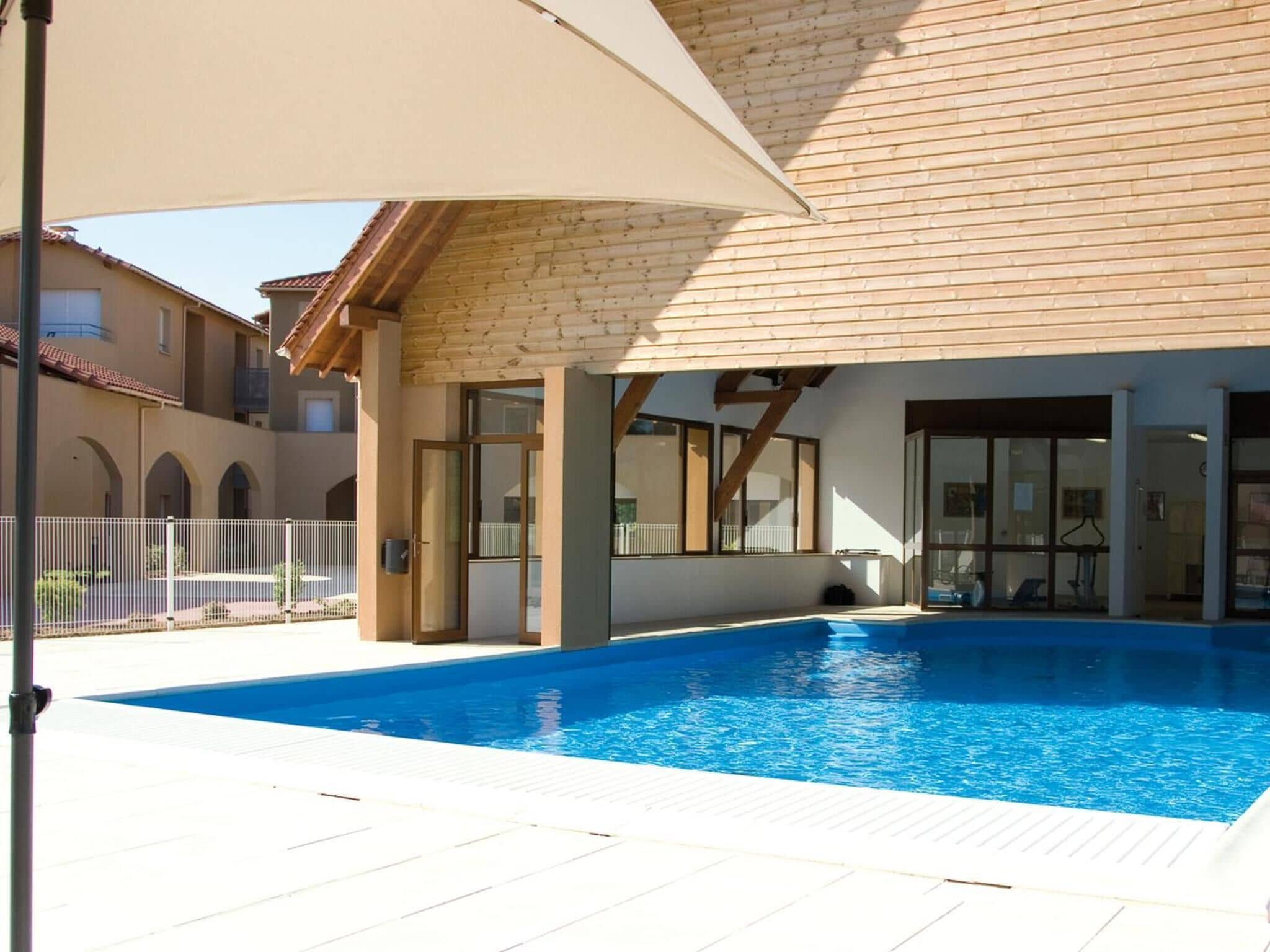 A swimming pool area with a covered patio, a wooden deck, and a building with large windows and a tiled roof.