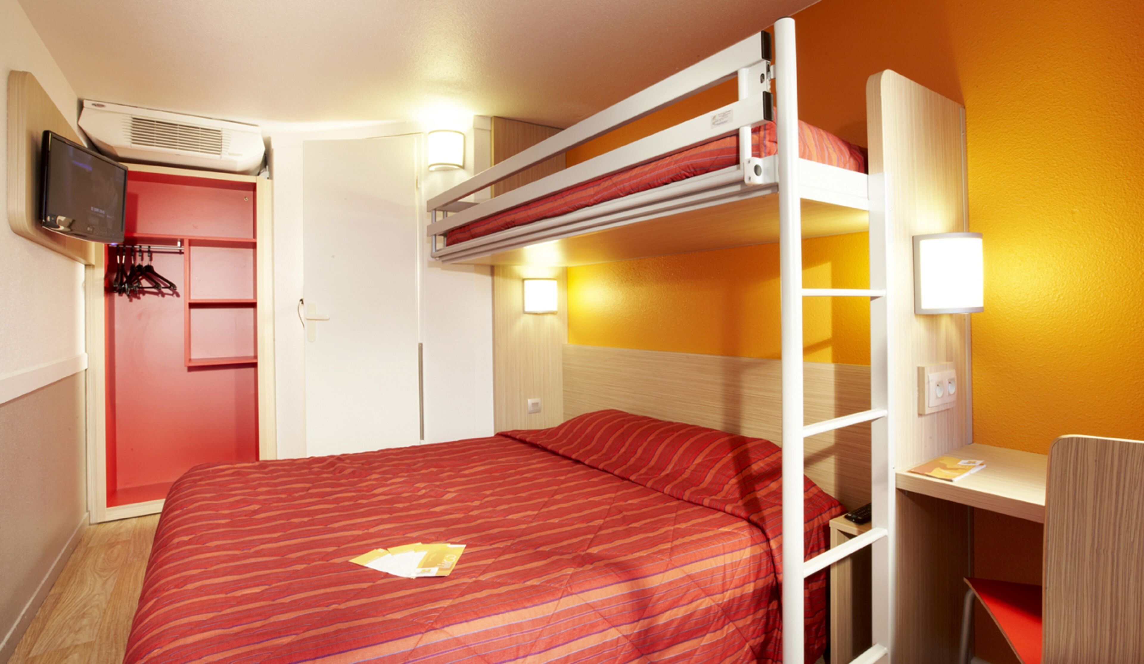 A bunk bed room with a red-striped bedspread, a TV mounted on the wall, and a small desk with a lamp.