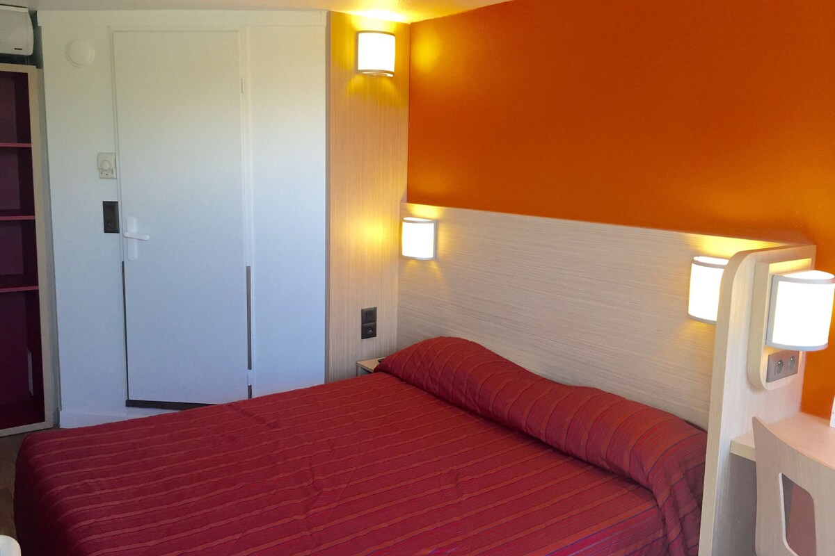 A hotel room with a bed, a door, and two wall-mounted lights.