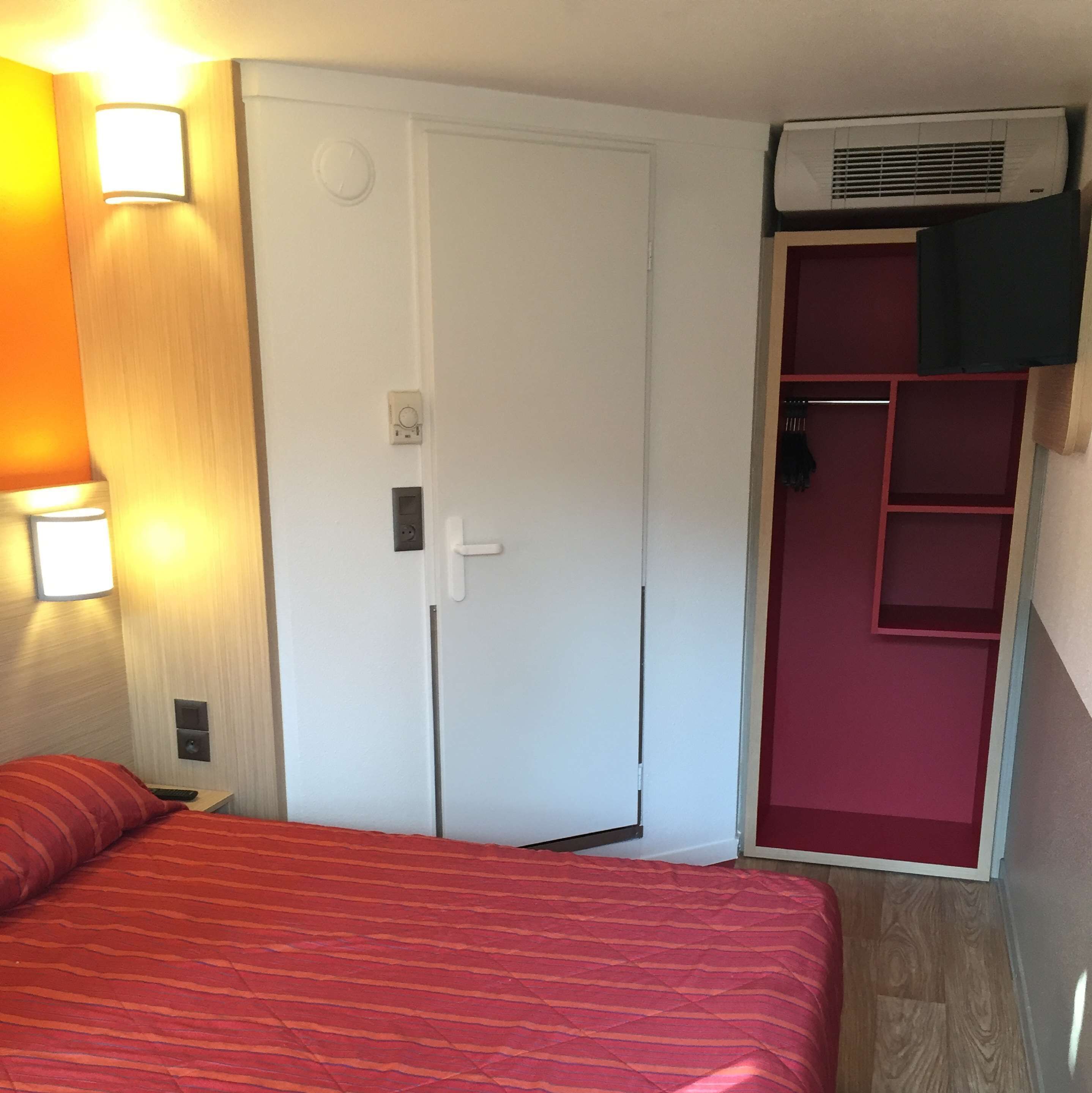 A hotel room with a bed, a wardrobe, a wall-mounted air conditioner, and a small shelf unit.