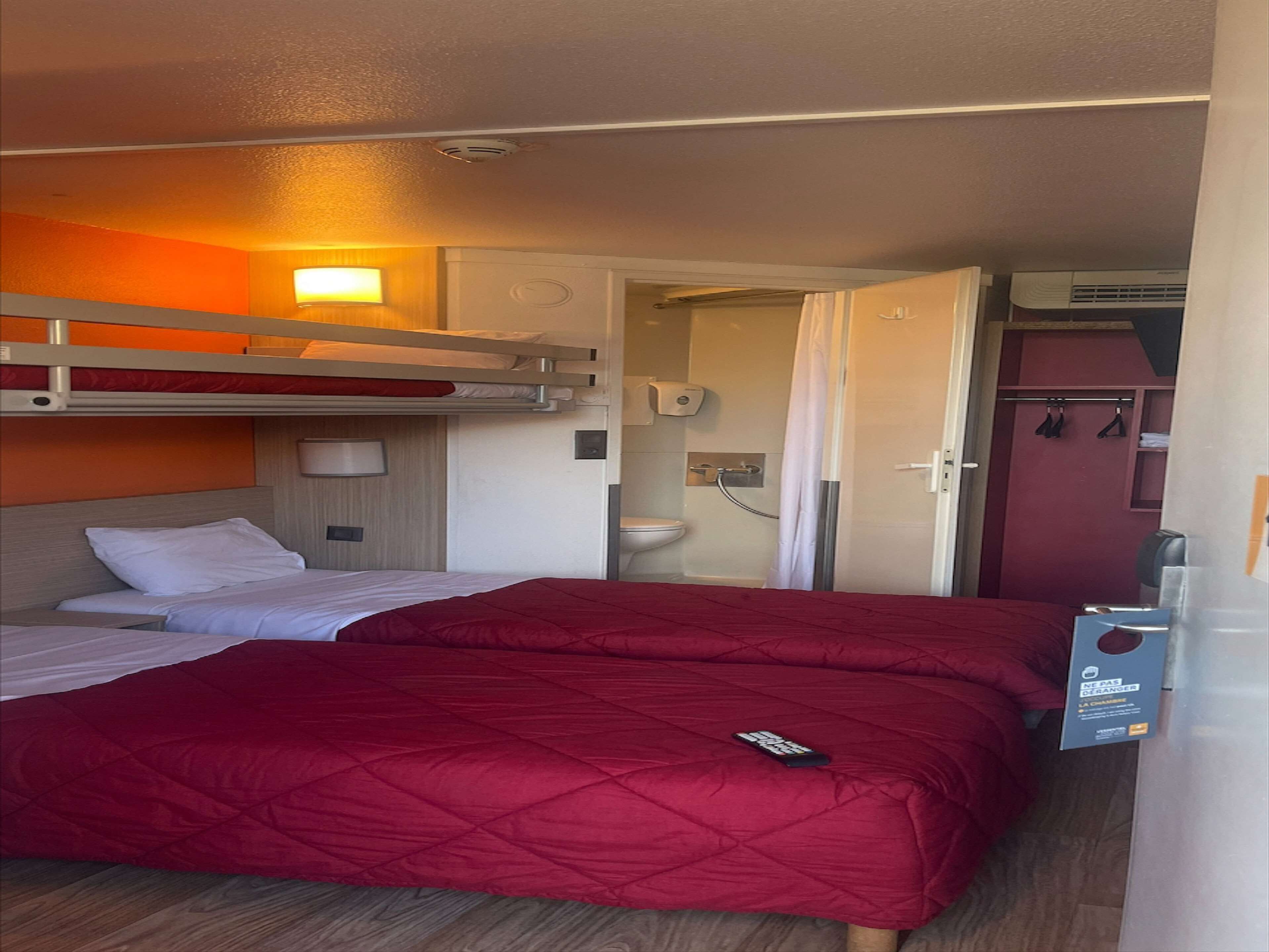 A compact hotel room with a bunk bed, a single bed with a red quilt, a bedside table, a lamp, a shower area, and a small closet.