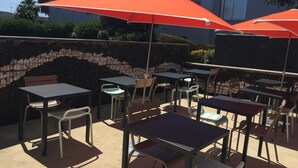 An outdoor patio with tables and chairs, some shaded by orange umbrellas, and a stone wall in the background. - Premiere Classe SETE - Balaruc (Balaruc-le-Vieux)