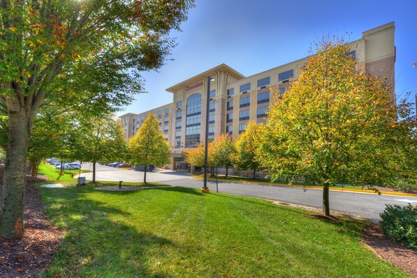 A hotel with a modern design, featuring a large parking lot and a well-maintained lawn with trees.
