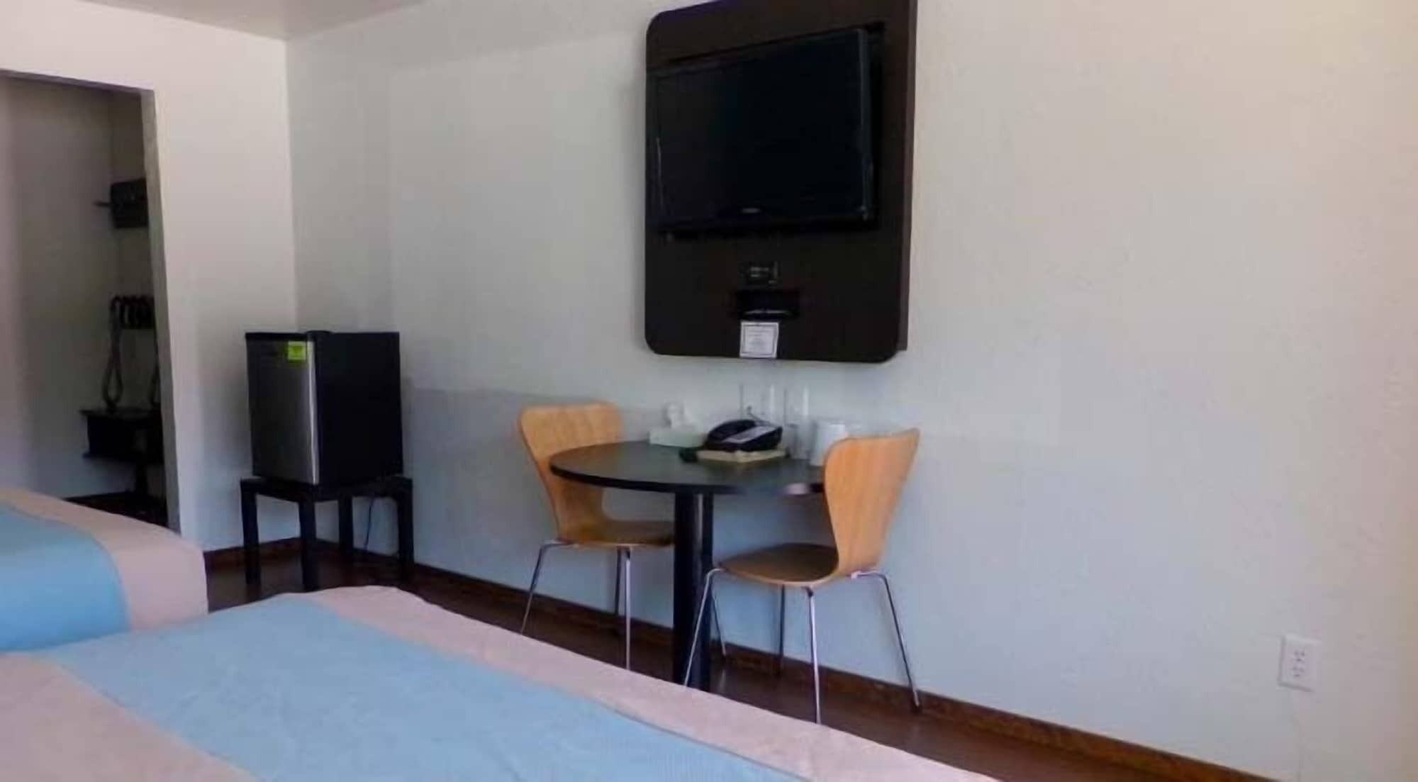 A hotel room with a bed, a desk with two chairs, a television mounted on the wall, and a small cabinet.