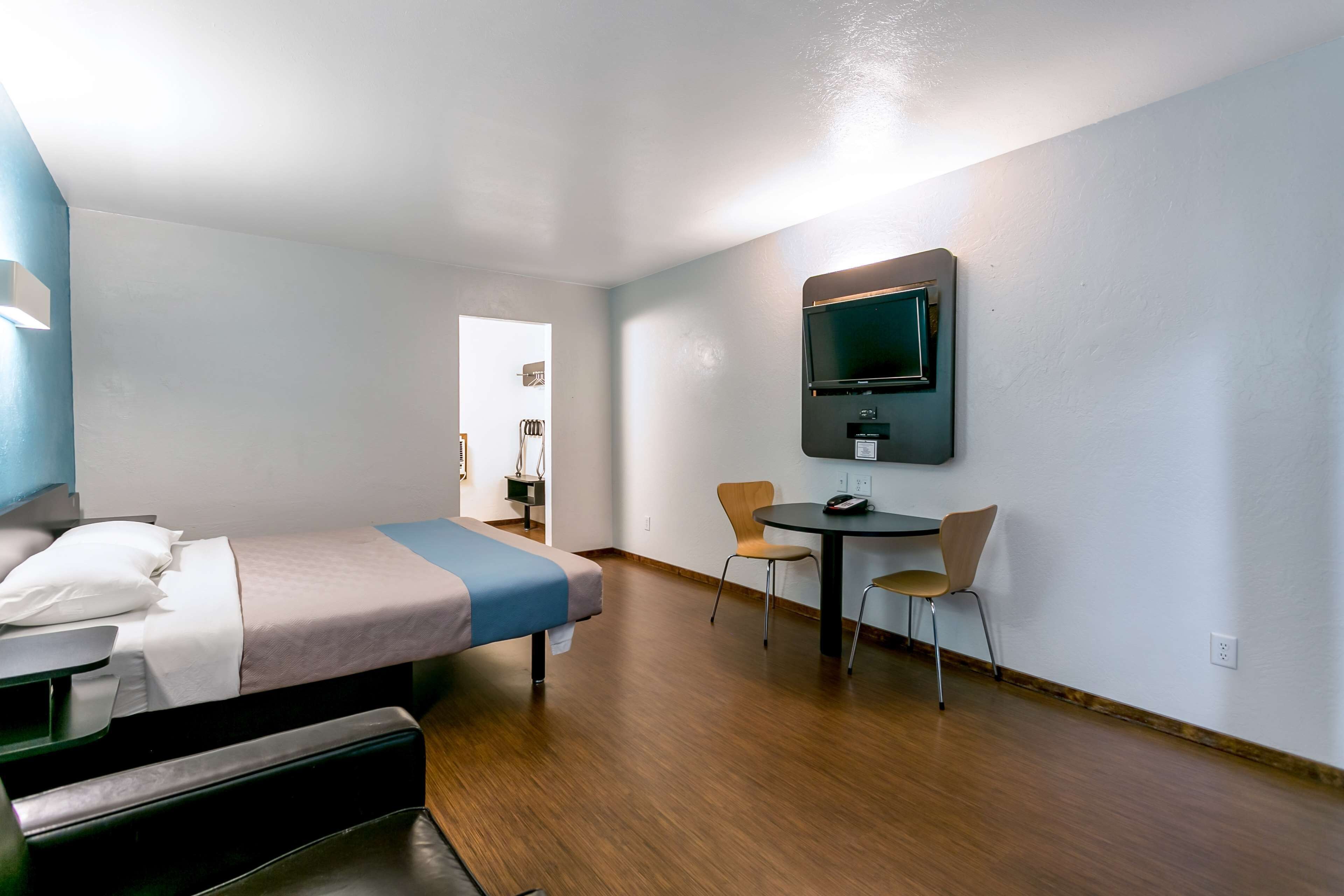 Una habitación de hotel con cama, escritorio, dos sillas, televisor montado en la pared y puerta que da a otra habitación.