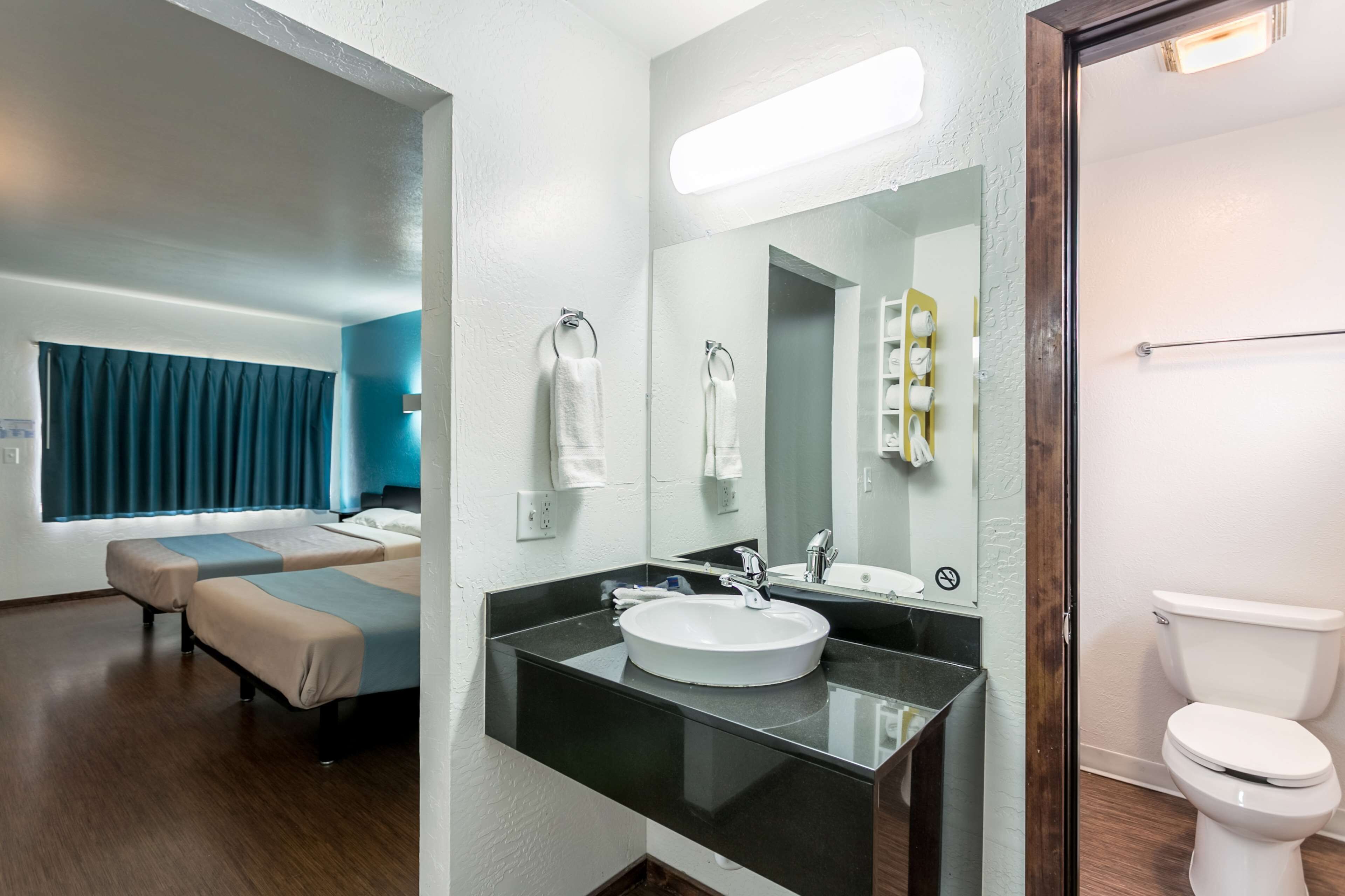 A modern bathroom with a black countertop, white sink, and a large mirror. There is a toilet and a bed with a blue curtain in the background.