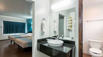 A modern bathroom with a black countertop, white sink, and a large mirror. There is a toilet and a bed with a blue curtain in the background.