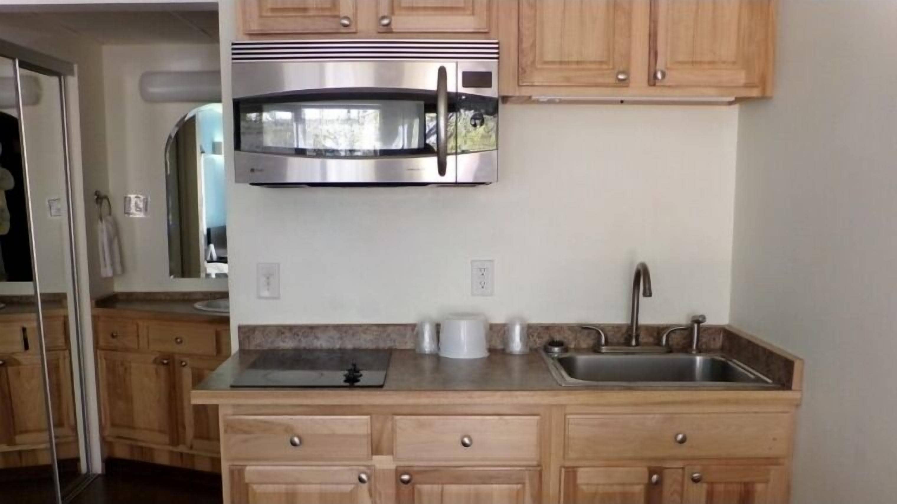 A kitchen with wooden cabinets, a stainless steel microwave, a sink, and a countertop with a kettle and cups.