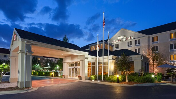 A hotel entrance with a covered driveway, flag poles, and a well-lit facade. - Hilton Garden Inn Gainesville (Gainesville)