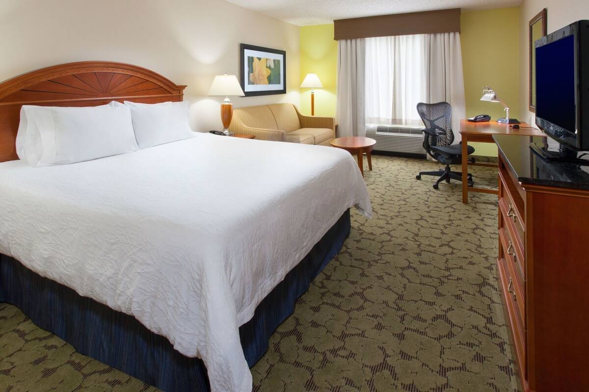 A hotel room with a large bed, a desk with a computer, a chair, a television, and a framed picture on the wall.
