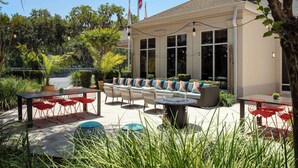 An outdoor seating area with tables and chairs, surrounded by greenery and a building with large windows. - Hilton Garden Inn Gainesville (Gainesville)