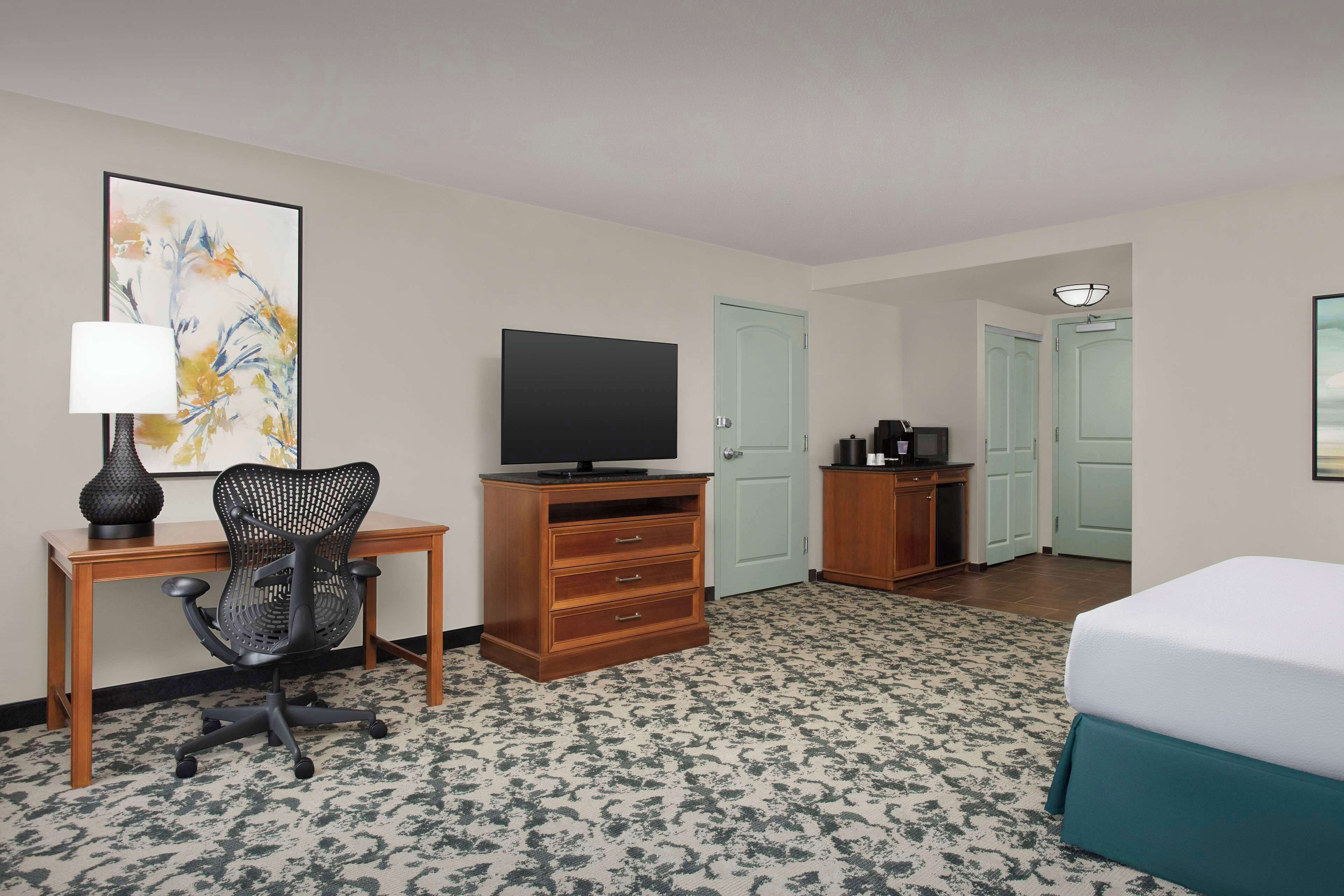 A hotel room with a bed, a desk with a chair, a television, a dresser, and a door.