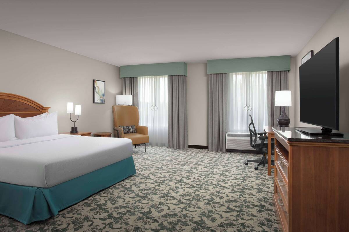 A hotel room with a large bed, a desk with a flat-screen TV, a chair, and a desk lamp.
