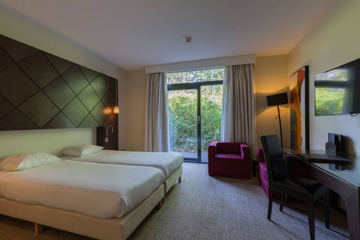 Habitación de hotel con cama, escritorio, silla, televisor y ventana con cortinas.