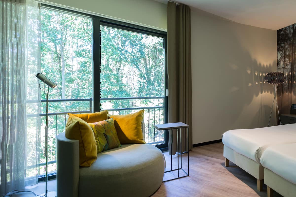 Una habitación de hotel con un ventanal grande, una cama, una mesita de noche y un cuadro de un bosque.