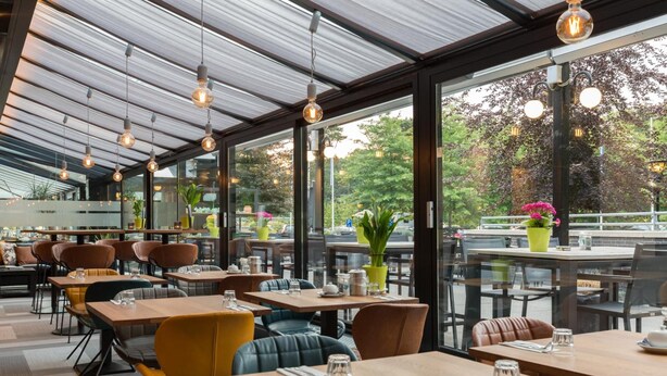 Un restaurant au décor moderne, avec des tables et des chaises en bois, de grandes fenêtres et des luminaires suspendus.