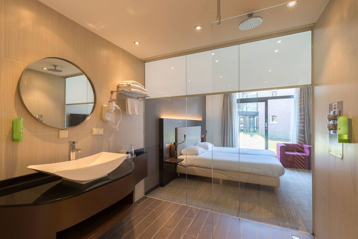 Habitación de hotel moderna con un espejo grande, una bañera, una cama y vistas al exterior.
