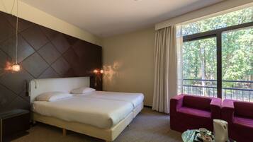 A hotel room with a large bed, a sofa, a coffee table, and a window with a view of trees.
