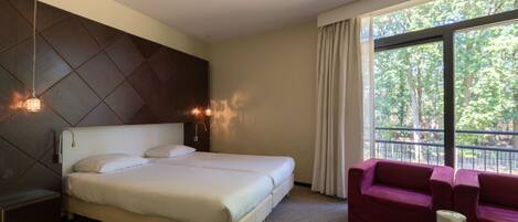 A hotel room with a large bed, a sofa, a coffee table, and a window with a view of trees.