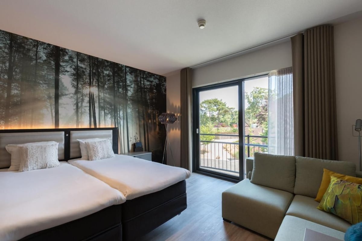 Ein Hotelzimmer mit einem großen Bett, einer Sitzecke, einem Waldbild und einem Balkon mit Blick auf Bäume.