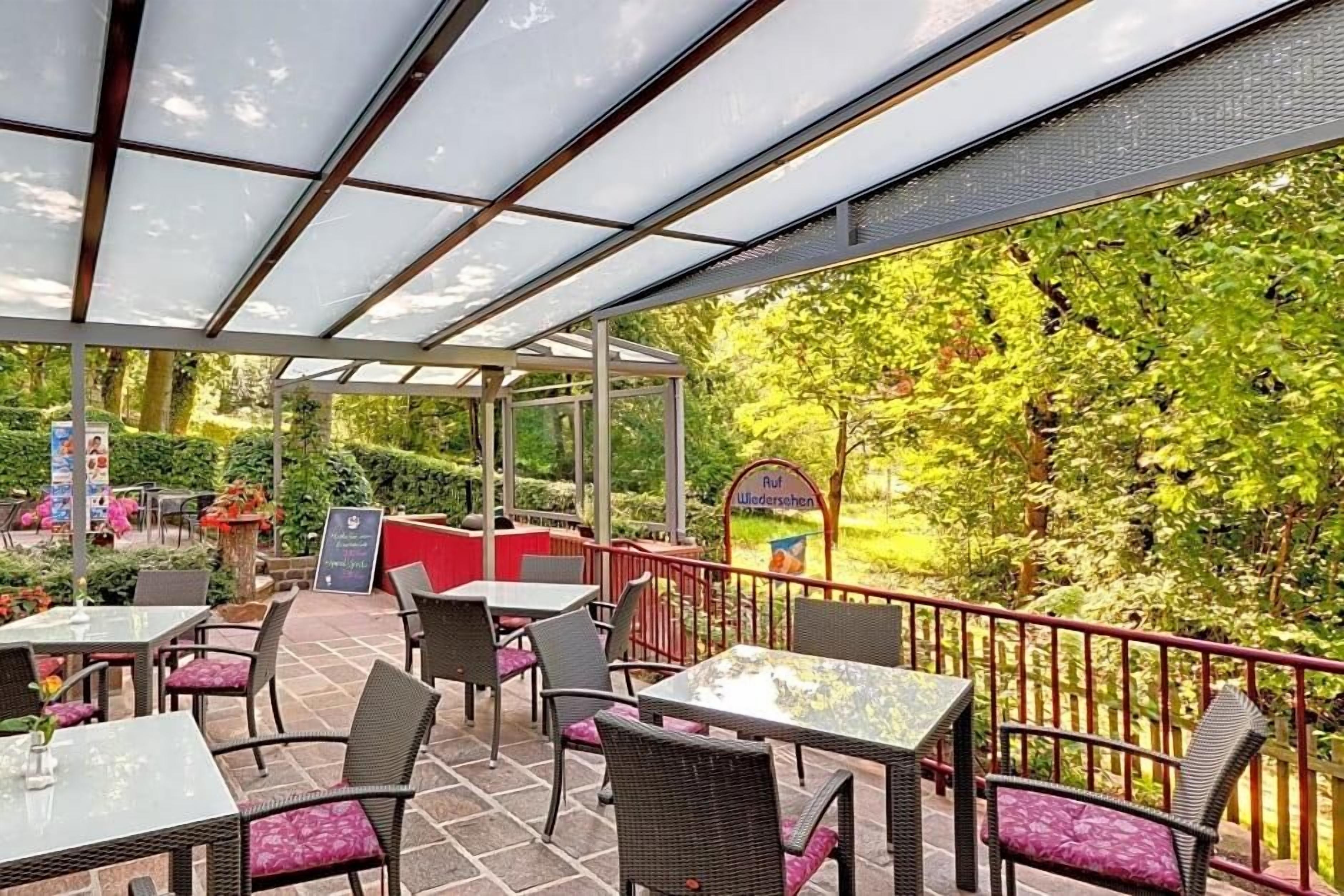 Un espace extérieur aménagé en terrasse, avec des tables et des chaises, entouré de verdure et protégé par une paroi de verre.