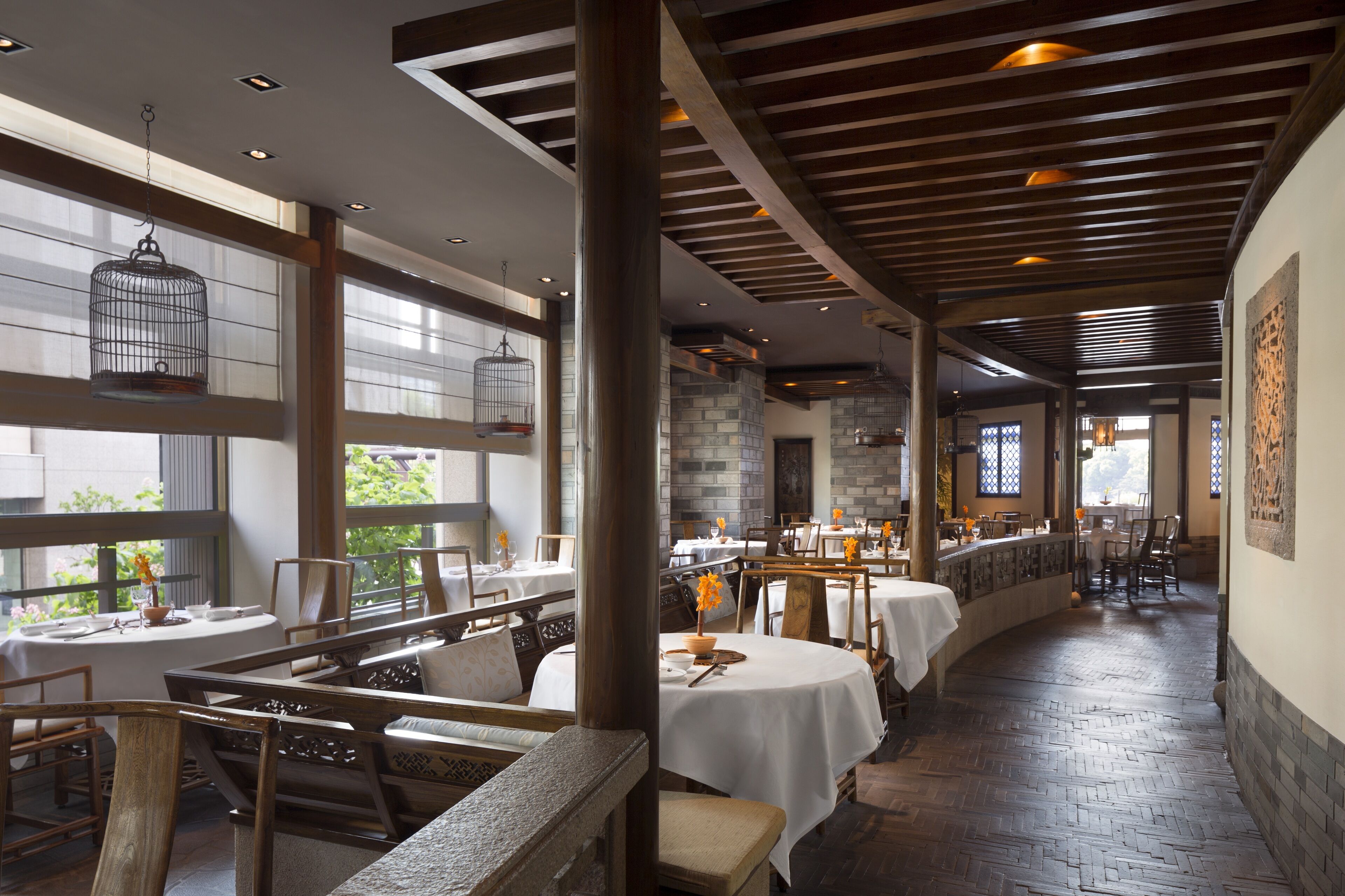 A restaurant with wooden beams, large windows, and a brick floor. Tables are set with white tablecloths and chairs.