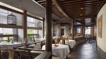 A restaurant with wooden beams, large windows, and a brick floor. Tables are set with white tablecloths and chairs.