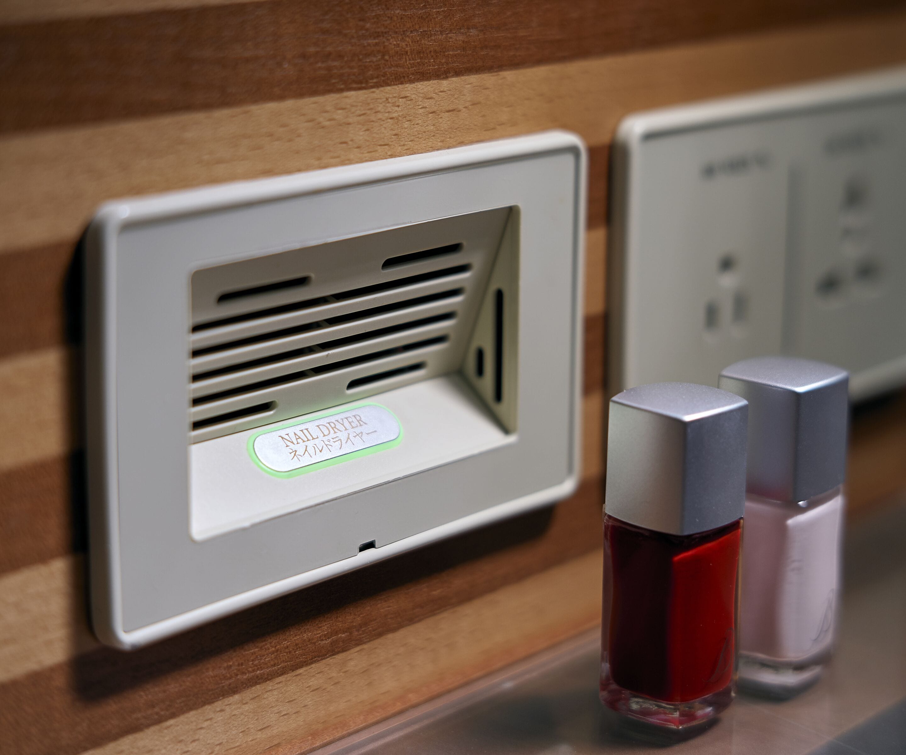 A wall-mounted device labeled "Nail Dryer" with two bottles of red and pink nail polish on a wooden surface.