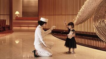 A person in a white uniform kneeling and interacting with a child in a black dress in a modern interior with wooden paneling and a large, circular architectural feature.